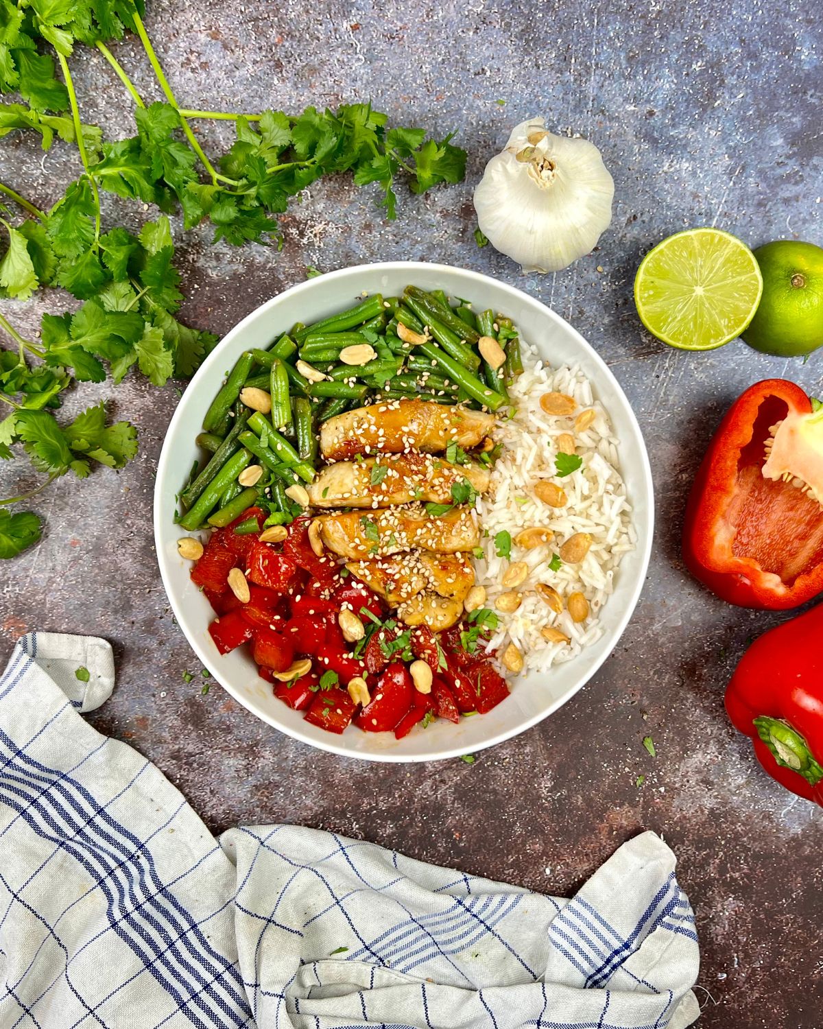 Eine Schüssel Reis mit Hähnchenstreifen, grünen Bohnen, Tomatenwürfeln, Erdnüssen und Sesamkörnern steht auf einer strukturierten Oberfläche. Um die Asia-Bowl mit grünen Bohnen herum befinden sich frischer Koriander, Knoblauch, Limettenhälften, eine rote Paprika und ein blau-weiß kariertes Tuch.