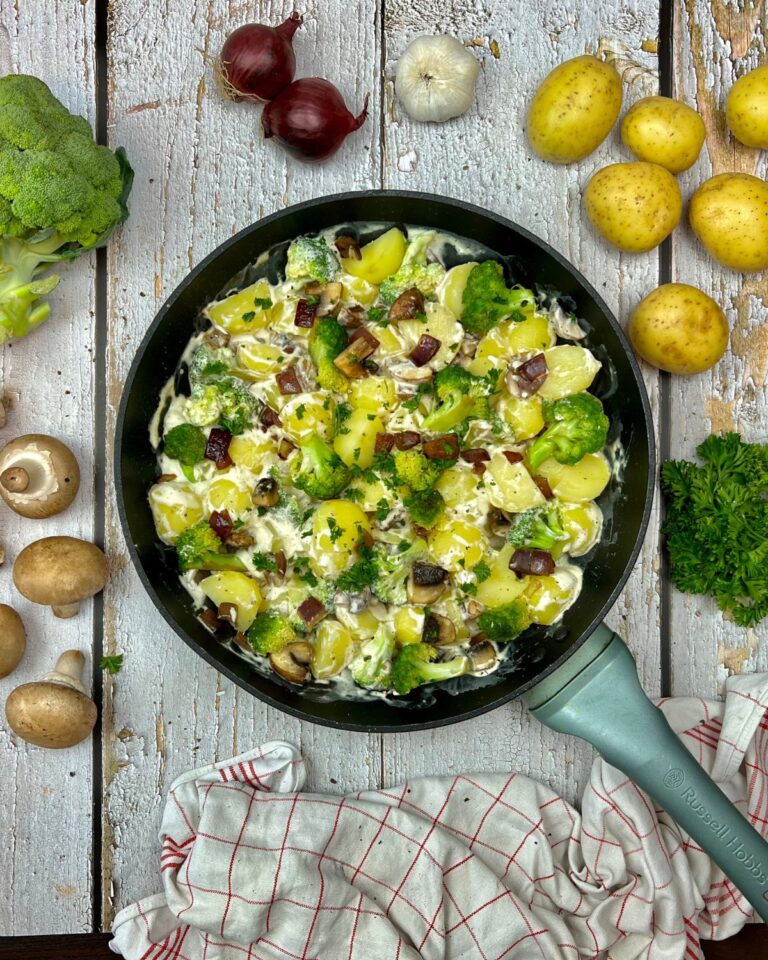 Eine Pfanne mit einer cremigen Brokkoli-Kartoffel-Pfanne, belegt mit Brokkoli, gewürfelten Champignons und frischer Petersilie steht auf einem rustikalen Holztisch. Um die Pfanne herum liegen Zutaten wie kleine Kartoffeln, rote Zwiebeln, Brokkoli, Pilze und ein rot-weißes Tuch.