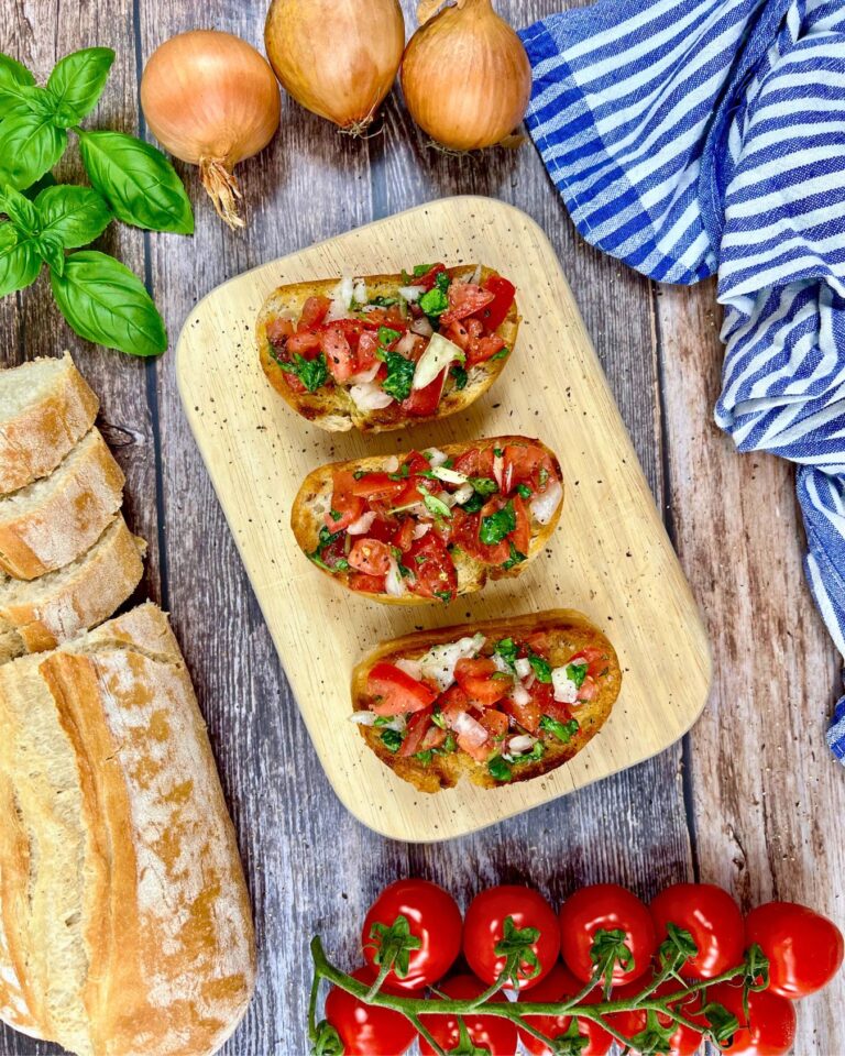 Draufsicht auf drei Bruschetta-Brote-Stücke, belegt mit gewürfelten Tomaten, Zwiebeln und Kräutern auf einer Holzplatte. Frisches Basilikum, Zwiebeln, ein gestreiftes Tuch, ein Laib Brot und Cherrytomaten umgeben sie auf einem rustikalen Holztisch.