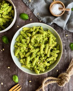 Eine Schüssel Fusilli-Nudeln ist mit leuchtend grüner Zucchinisauce überzogen und mit Pinienkernen garniert. Die Schüssel steht auf einem rustikalen Holztisch, daneben liegen Basilikumblätter, ein Schälchen mit grobem Salz und ein Holzlöffel. Neben der Stoffserviette liegt ein weiterer Teller mit cremigen Nudeln mit Zucchinisauce.