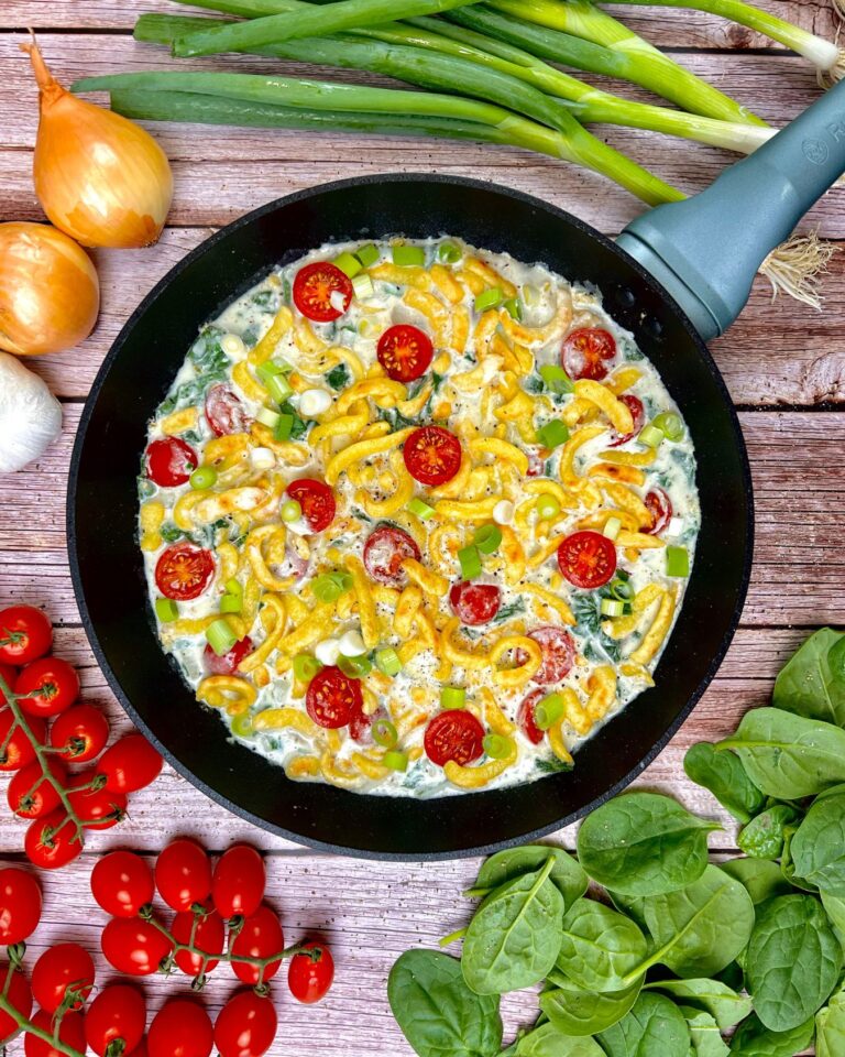 Eine cremige Spätzle Pfanne mit Spinat und Tomaten. Umgeben von frischen Zutaten – Kirschtomaten, Zwiebeln, Frühlingszwiebeln, Spinat und Knoblauch – ruht es auf einer rustikalen Holzoberfläche.