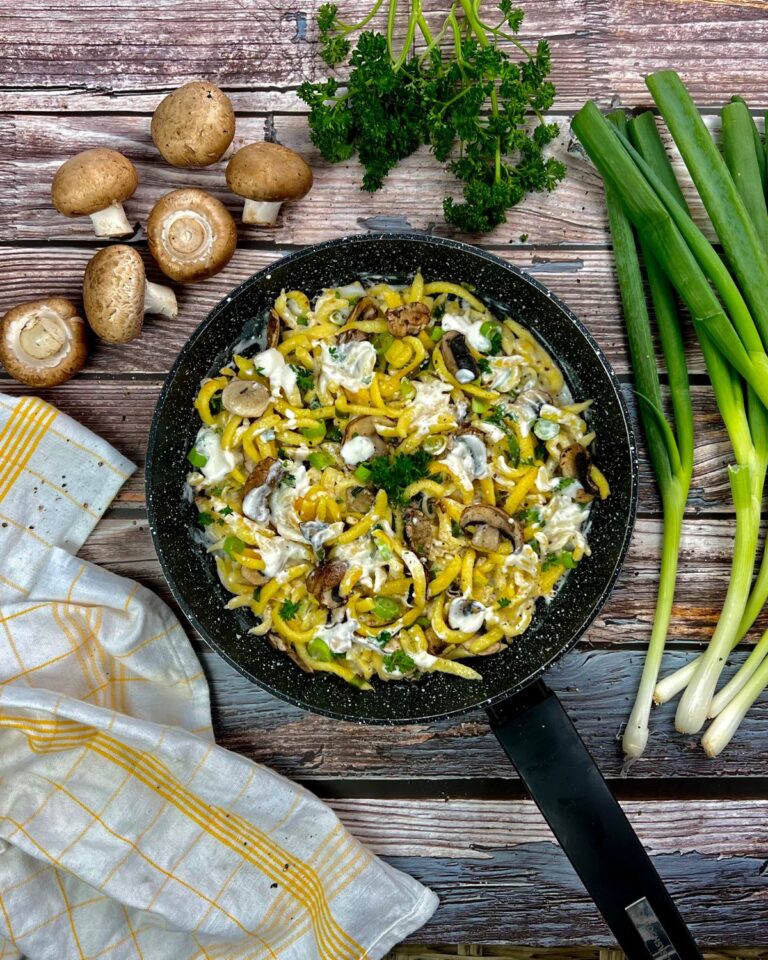 Eine Pfanne mit einer cremigen Spätzlepfanne, garniert mit frischer Petersilie, steht auf einer rustikalen Holzoberfläche. Um die Pfanne herum liegen ganze Champignons, Frühlingszwiebeln und ein gelb-weiß kariertes Handtuch. Diese cremige Spätzlepfanne mit Champignons bietet eine einladende Geschmacksmischung.