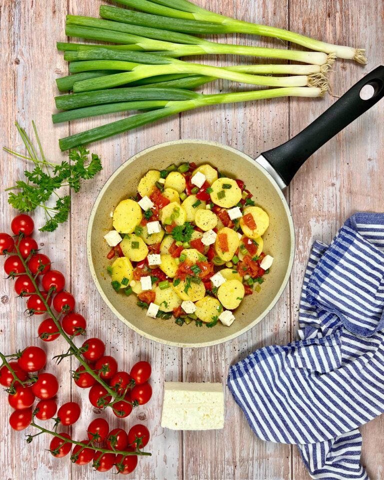 Neben dieser einfachen Kartoffelpfanne mit Cherrytomaten und Feta liegen ganze Frühlingszwiebeln, Cherrytomaten am Strauch, ein Stück Käse, Petersilie und ein gestreiftes Tuch auf einer Holzoberfläche.