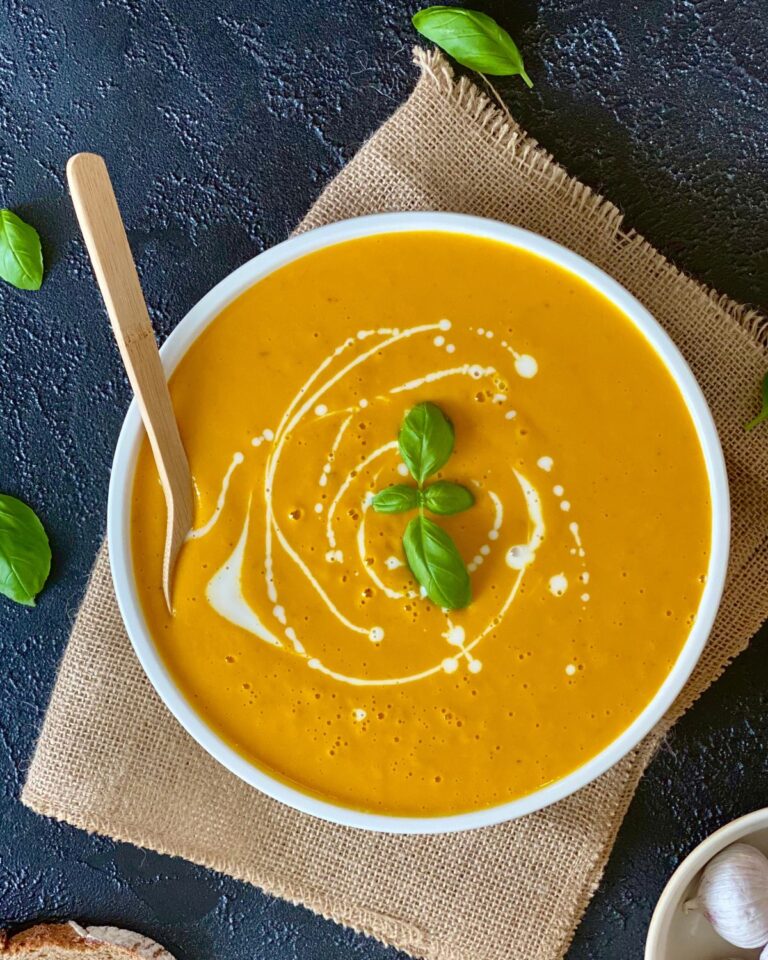 Eine Schüssel cremige Kürbissuppe aus dem Ofen, garniert mit einem Klecks Sahne und frischen Basilikumblättern, in der Schüssel liegt ein Holzlöffel. Die Schüssel steht auf einem Stück Sackleinen auf einer dunklen, strukturierten Oberfläche, auf dem Basilikumblätter verstreut sind.