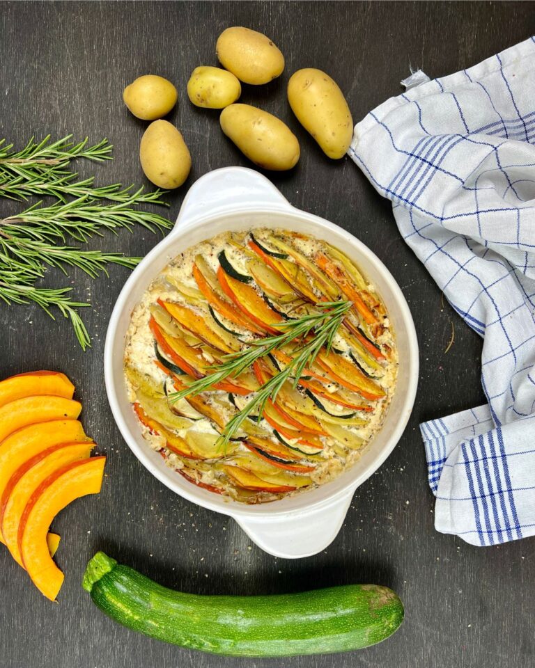 Eine runde, weiße Keramik-Auflaufform, gefüllt mit einem Kürbis-Kartoffel-Auflauf, belegt mit kreisförmig angeordneten Zucchini-, Kürbis- und roten Kartoffelscheiben. Um die Form herum liegen Zweige frischen Rosmarins, ganze Kartoffeln, eine Zucchini, zwei Kürbisstücke und ein gestreiftes Tuch auf einer dunklen Oberfläche.