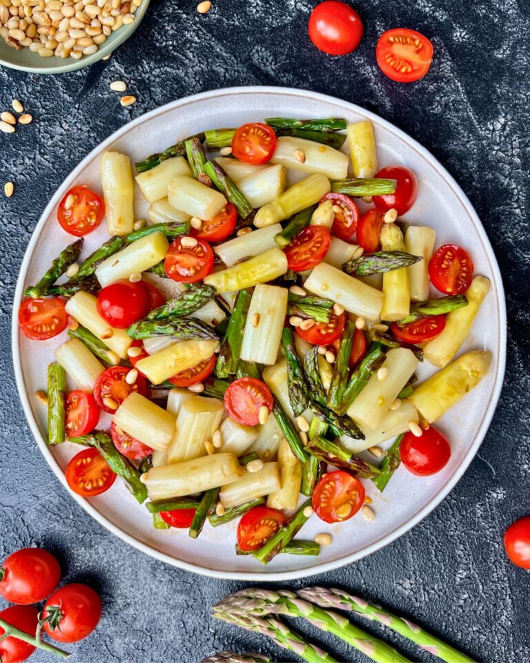 Ein Teller Spargelsalat mit Cherrytomaten und Pinienkernen präsentiert weißen und grünen Spargel, Cherrytomaten und Pinienkerne auf einem dunklen strukturierten Hintergrund.