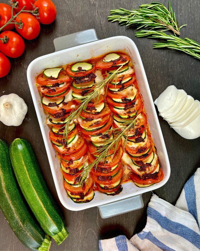 Ein gebackener Tomaten-Zucchini-Auflauf mit Mozzarella besteht aus geschnittenen Zucchini, Cherrytomaten und Käse, garniert mit Rosmarin in einer weißen Auflaufform.