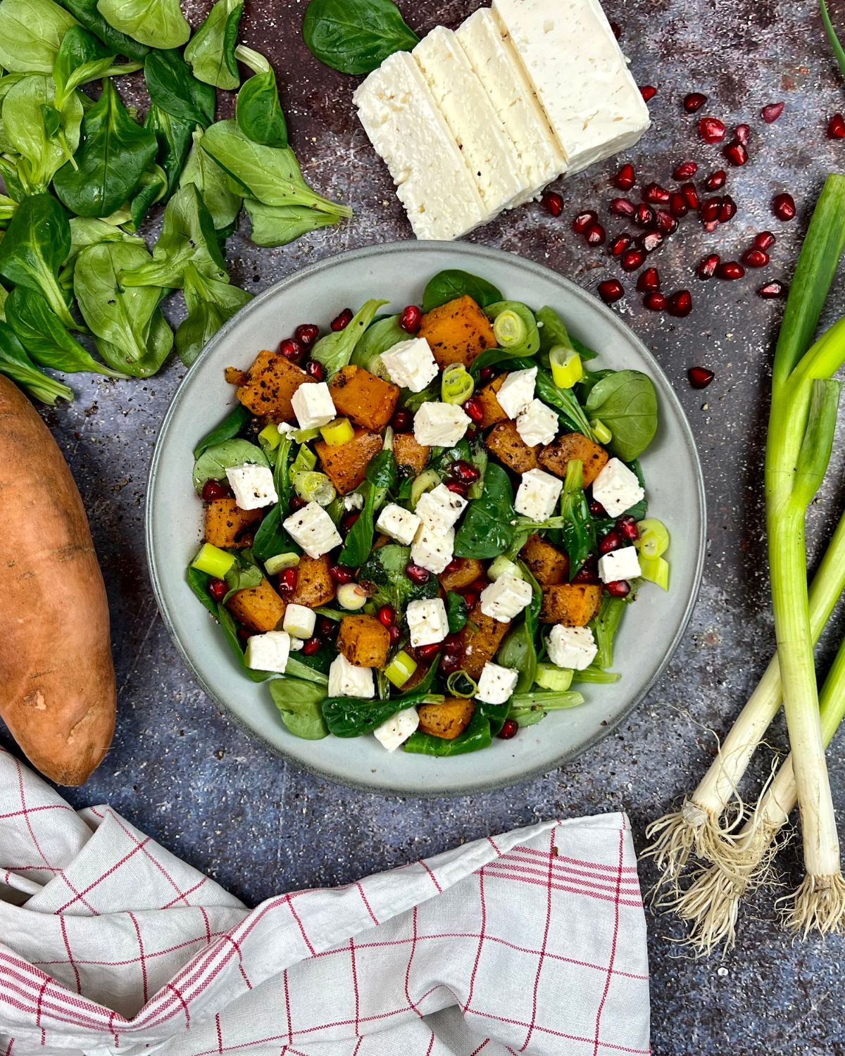 Auf einem grauen Teller wird ein frischer Salat präsentiert, der geröstete Süßkartoffeln, Blattgemüse, gewürfelten Fetakäse, Granatapfelkerne und gehackte Frühlingszwiebeln enthält. Zu den umliegenden Zutaten gehören Spinatblätter, Frühlingszwiebeln, eine Süßkartoffel, Fetablöcke und ein rotkariertes Tuch. Dieser köstliche Feldsalat mit Süßkartoffeln bietet eine lebendige und leckere Note.