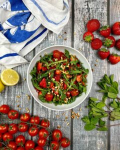 Eine weiße Schüssel mit Erdbeer-Rucola-Salat, garniert mit Pinienkernen, steht auf einer rustikalen Holzoberfläche. Um die Schüssel herum befinden sich frische Erdbeeren, Kirschtomaten an der Rispe, Rucola, eine halbierte Zitrone und ein blau-weißes Tuch.