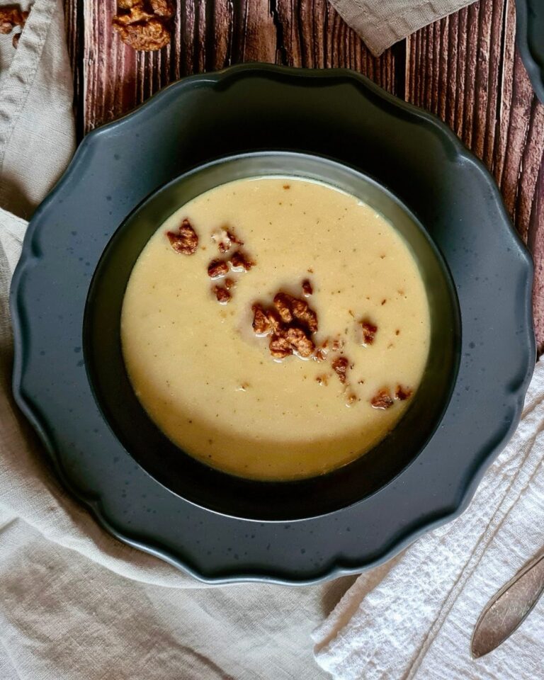 Eine schwarze Schüssel gefüllt mit cremiger gelber Chicoreesuppe steht auf einer Holzfläche. Braune, knusprige Toppings sind auf der Suppenoberfläche verstreut. Neben der Schüssel liegen ein helles Tuch und ein teilweise sichtbarer Löffel.