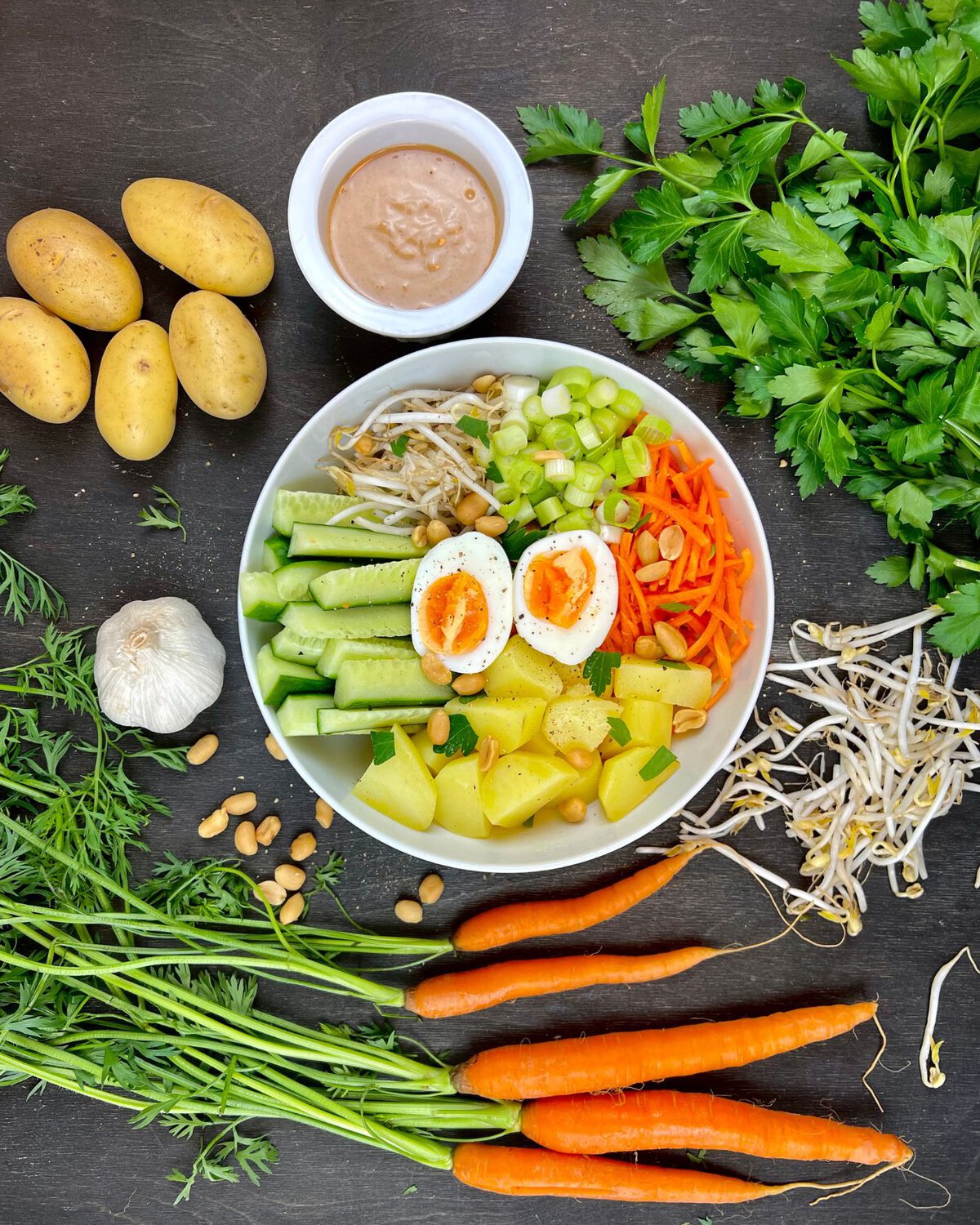 Im Mittelpunkt steht eine farbenfrohe Schüssel Gado-Gado, umgeben von frischen Kartoffeln, Petersilie, Knoblauch, Karotten und knackigen Sojasprossen. In der Schüssel befinden sich gekochte Eier, Gurken, gehaltvolle Erdnüsse und zarte Kartoffeln, perfekt ergänzt durch eine kleine Schale pikanter Erdnusssauce.