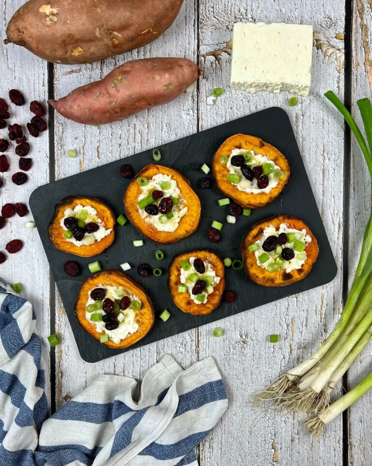 Auf einem rustikalen Holztisch steht eine schwarze Schieferplatte mit sechs gebackenen Süßkartoffel-Scheiben, belegt mit cremigem Käse, gehackten Frühlingszwiebeln, Preiselbeeren und Blaubeeren. Um die Platte herum liegen ganze Süßkartoffeln, ein Stück Käse, Frühlingszwiebeln und ein Küchentuch.