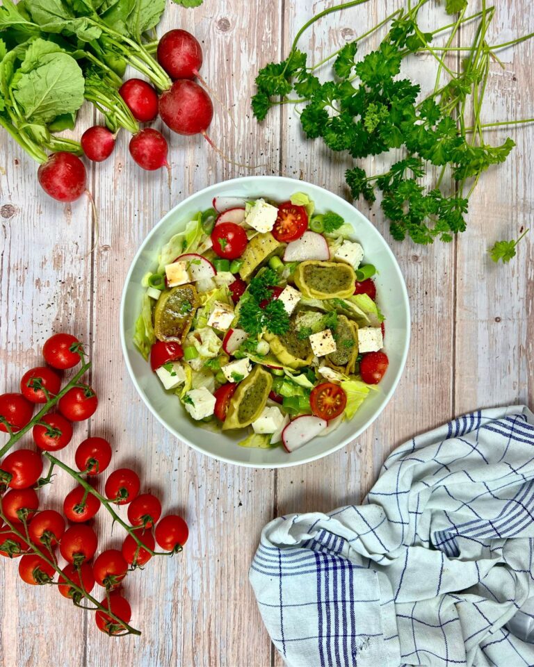 Ein frischer Gesunder Maultaschen-Salat mit Salat, Kirschtomaten, Radieschen und Fetakäse garniert mit Petersilie. Um die Schüssel herum liegen ganze Radieschen, Kirschtomaten am Strauch, Petersilie und ein kariertes Tuch auf einem Holztisch.