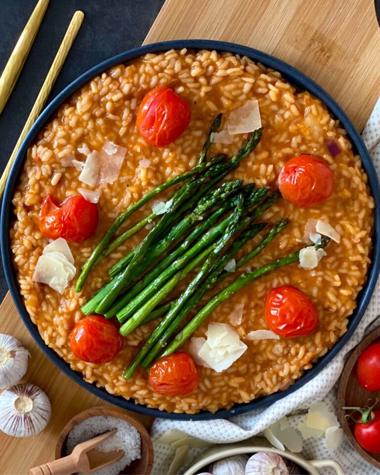 Ein Teller mit gesundem Tomaten-Risotto mit grünem Spargel, garniert mit Cherrytomaten und Parmesanspänen, steht zwischen Knoblauchknollen und Kochzutaten auf einer Holzoberfläche.