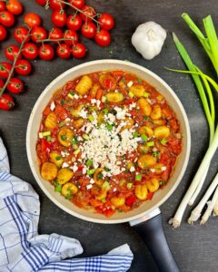 Eine Pfanne gefüllt mit Gnocchi mit Feta und Tomaten, garniert mit geriebenem Käse und gehackten Frühlingszwiebeln. Um die Pfanne herum sind Kirschtomaten an der Rebe, eine Knolle Knoblauch, frische Frühlingszwiebeln und ein blau-weiß kariertes Küchentuch.