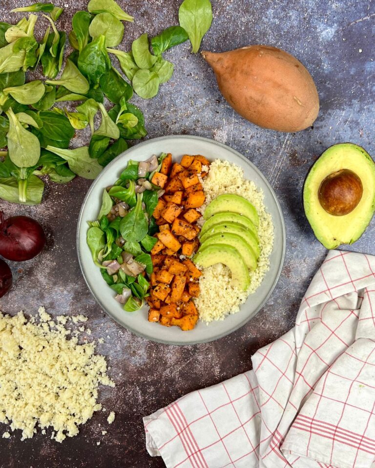 Eine Schüssel Quinoa, gefüllt mit geschnittener Avocado, gerösteten Süßkartoffeln und Blattgemüse, garniert mit gewürfelten roten Zwiebeln. Um die Schüssel herum liegen frische Spinatblätter, eine ganze Süßkartoffel, eine Avocado, rote Zwiebeln, eine Stoffserviette und etwas verstreutes Quinoa. Dieses Gericht erinnert an eine köstliche Couscous-Bowl mit Süßkartoffeln.