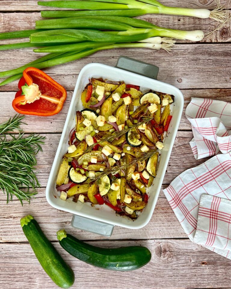 Ein Kartoffel-Zucchini-Auflauf mit Feta, darunter Zucchini, rote Paprika und Kartoffeln auf einem Holztisch. Um die Auflaufform herum liegen frische Frühlingszwiebeln, eine halbierte rote Paprika, Rosmarinzweige, eine ganze Zucchini und ein gestreiftes Tuch.