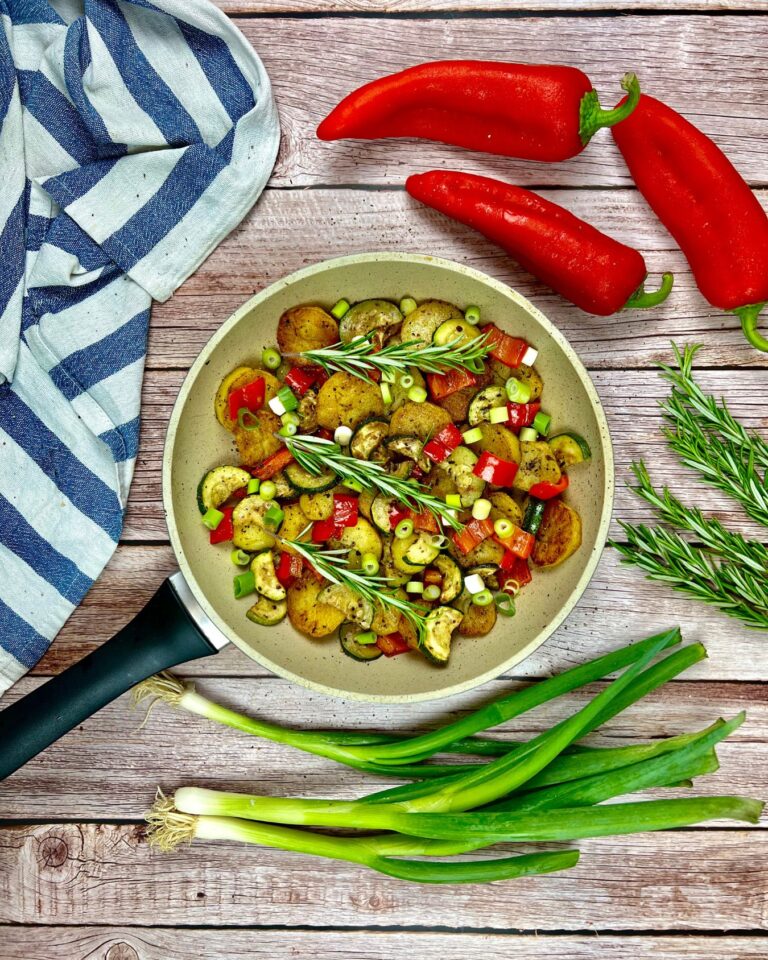 Eine Bratpfanne auf einem Holztisch, randvoll mit einer Kartoffel-Zucchini-Pfanne, in der gekochte Kartoffeln, Zucchini und rote Paprikaschoten, alles mit Rosmarin garniert, liegen.