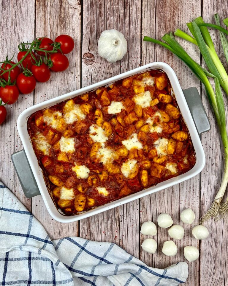 Ein Kürbis-Gnocchi-Auflauf, mit geschmolzenem Mozzarella-Käse in einer rechteckigen Keramik-Auflaufform. Um das Gericht herum befinden sich Zutaten: Cherrytomaten, eine Knoblauchknolle, Frühlingszwiebeln, Mini-Mozzarellakugeln und ein kariertes Geschirrtuch auf einer Holzoberfläche.