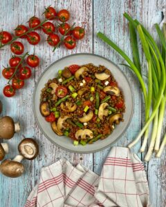 Eine Schüssel Linsen-Pilz-Ragout mit Kirschtomaten und Frühlingszwiebeln, auf einer rustikalen Holzoberfläche. Frische Cherrytomaten, Bund Frühlingszwiebeln, ganze Pilze und ein gestreiftes Tuch sind um die Schüssel herum angeordnet und erinnern an ein herzhaftes Linsen-Pilz-Ragout.