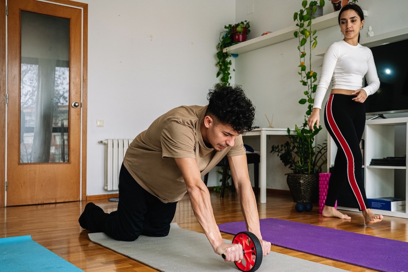 Mann benutzt Bauchmuskelroller waehrend Heimtraining Ein Mann kniet auf einer Yogamatte und benutzt einen Bauchmuskelroller.