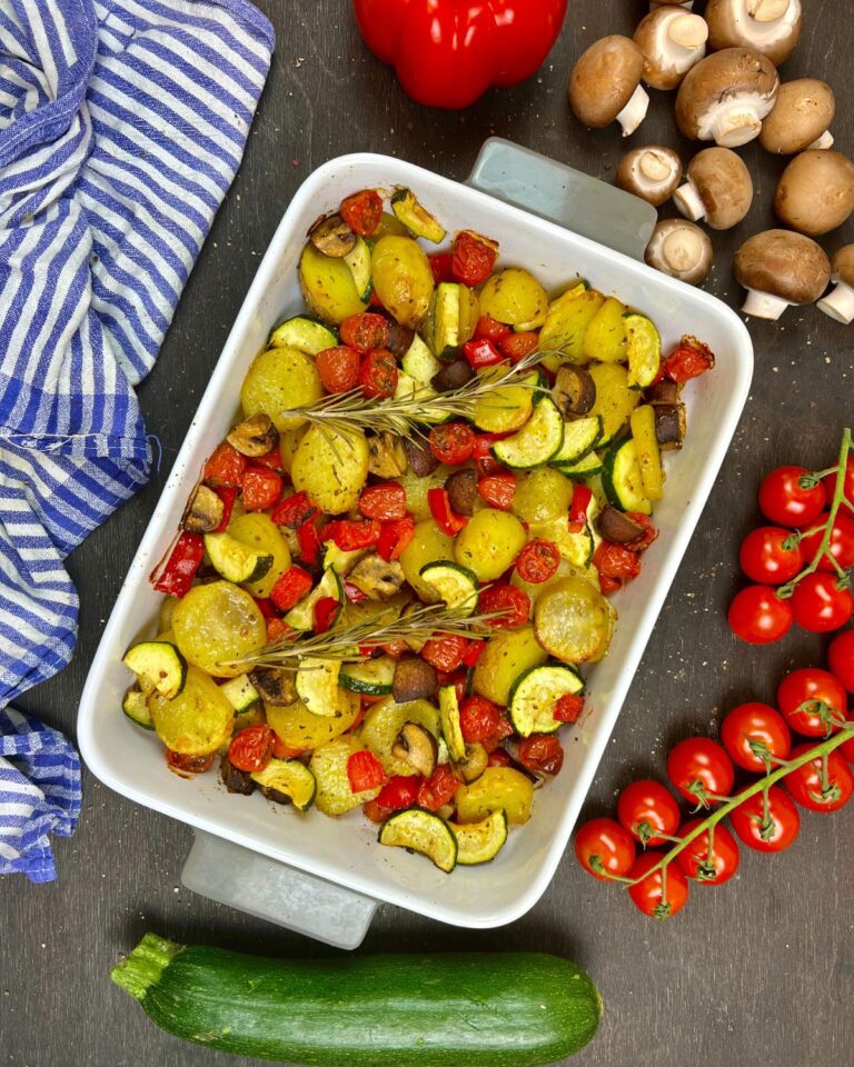 Eine weiße Auflaufform gefüllt mit mediterranem Ofengemüse, darauf Röstkartoffeln, Kirschtomaten, Zucchini, rote Paprika und Champignons, garniert mit Rosmarin. Um die Form herum liegen frische Zutaten: Kirschtomaten, Champignons, eine ganze Zucchini und eine rote Paprika. Auf der linken Seite liegt ein blau gestreiftes Tuch.