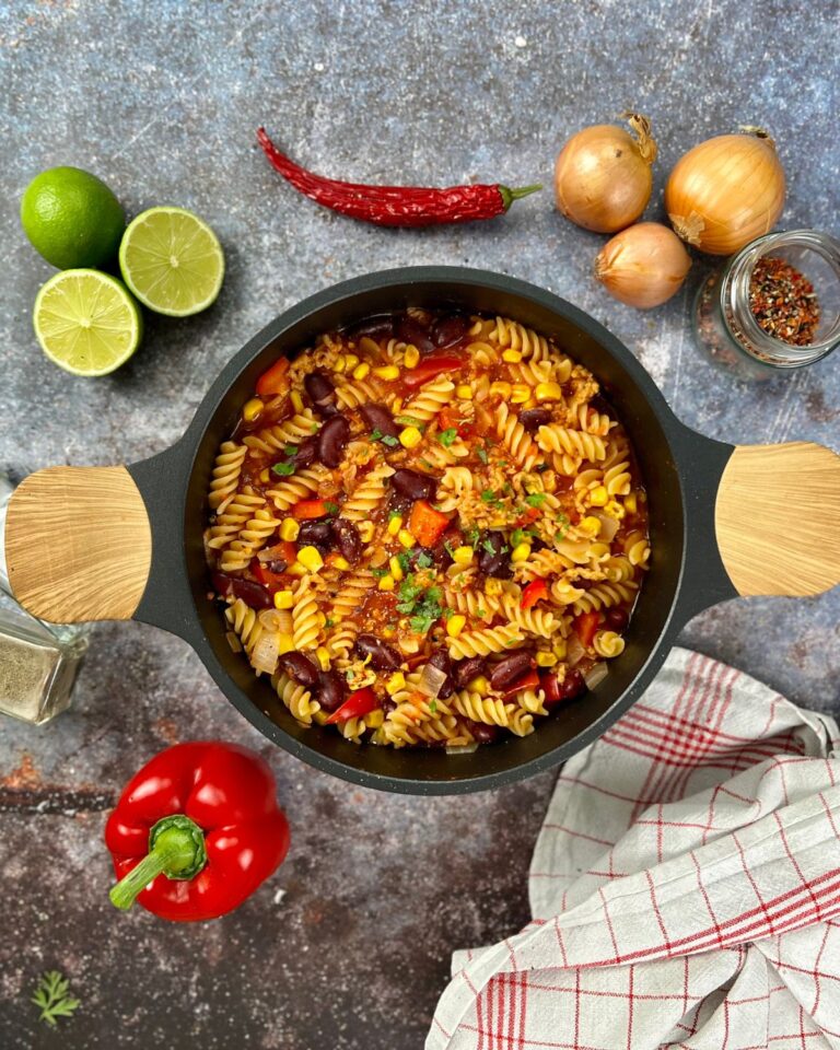 Ein schwarzer Topf mit einer mexikanischen One-Pot-Pasta aus Fusilli, Mais, Bohnen, roten Paprikaschoten und gehacktem Grünzeug steht auf einer strukturierten Oberfläche. Um den Topf herum sind Zutaten: Limettenhälften, eine rote Chilischote, Zwiebeln, rote Paprikaschoten und ein Glas Chiliflocken. Daneben steht ein rot-weiß kariertes Tuch.