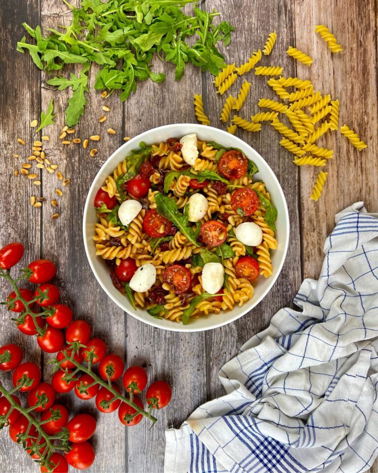 Eine Schüssel Fusilli-Nudelsalat mit Pesto, Cherrytomaten, Mozzarella-Kugeln, getrockneten Tomaten, Rucola und Pinienkernen steht auf einem rustikalen Holztisch.