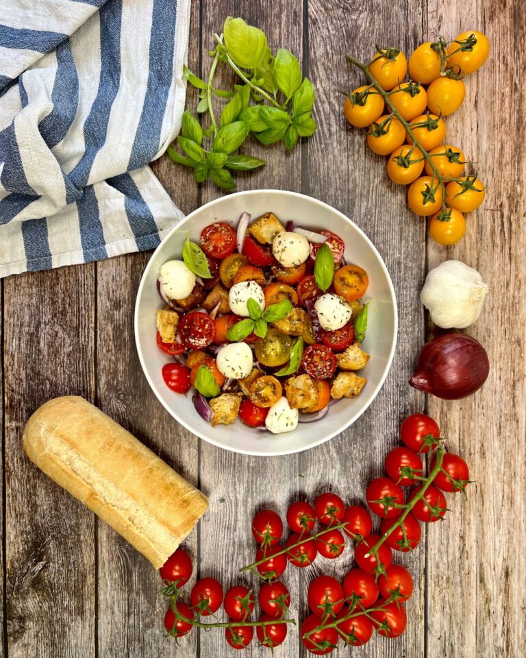 Auf einer Holzplatte steht eine Schüssel Caprese-Salat nach Panzanella-Art mit Kirschtomaten, Mozzarellakugeln, Basilikum und Croutons.