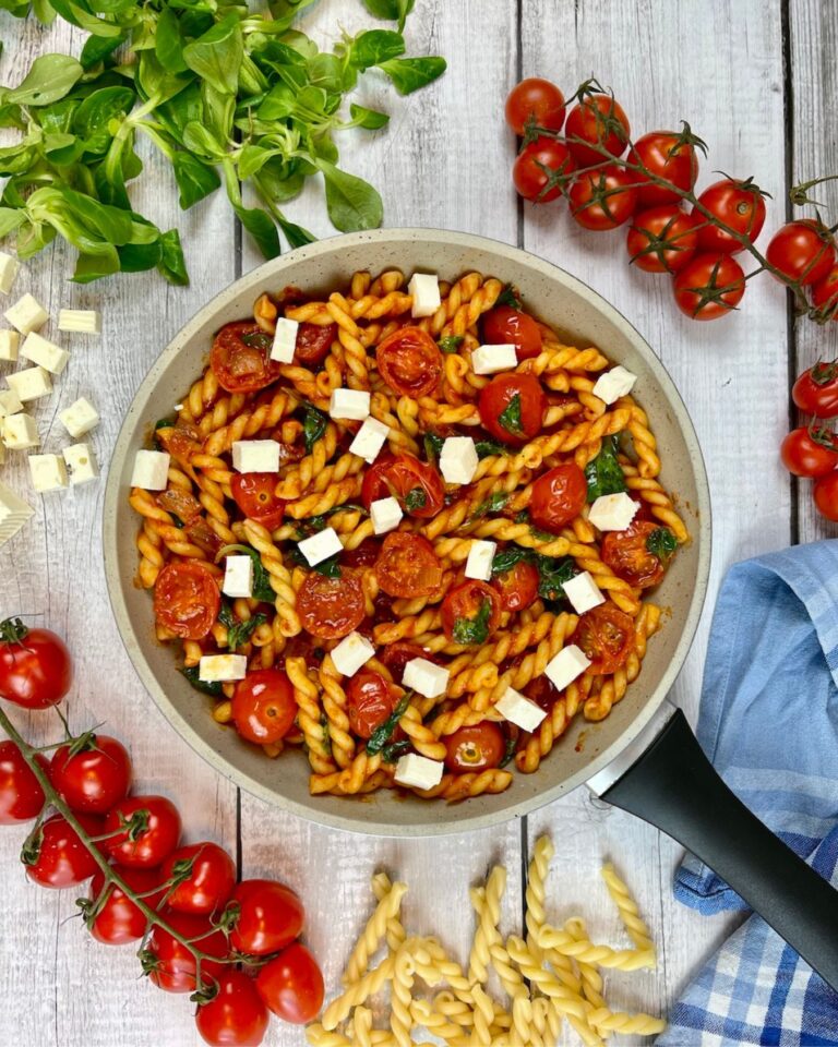 Auf einer Holzplatte steht eine Pfanne Pasta mit Tomaten und Feta.