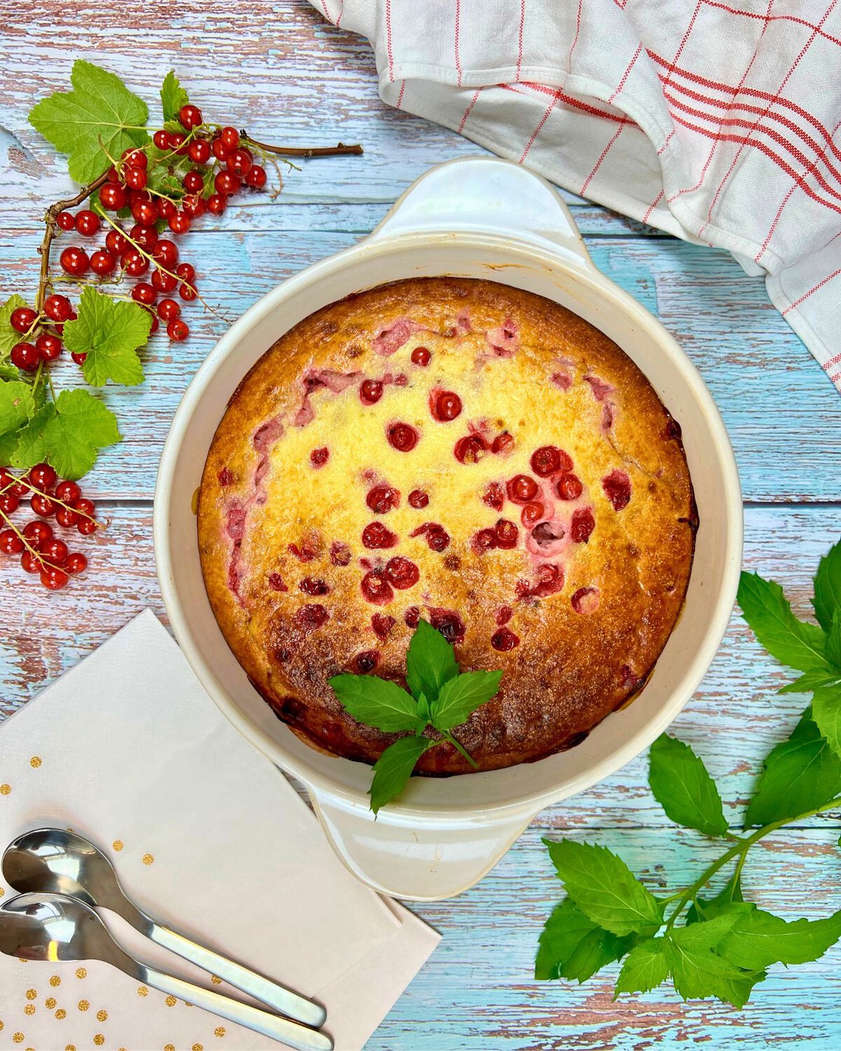 Ein proteinreicher Grießauflauf mit Johannisbeeren steht elegant in einer weißen Schale auf dem blauen Holztisch. Frische Johannisbeerzweige und grüne Blätter begleiten ihn, neben einem Tuch, einer weißen Serviette und zwei silbernen Löffeln.