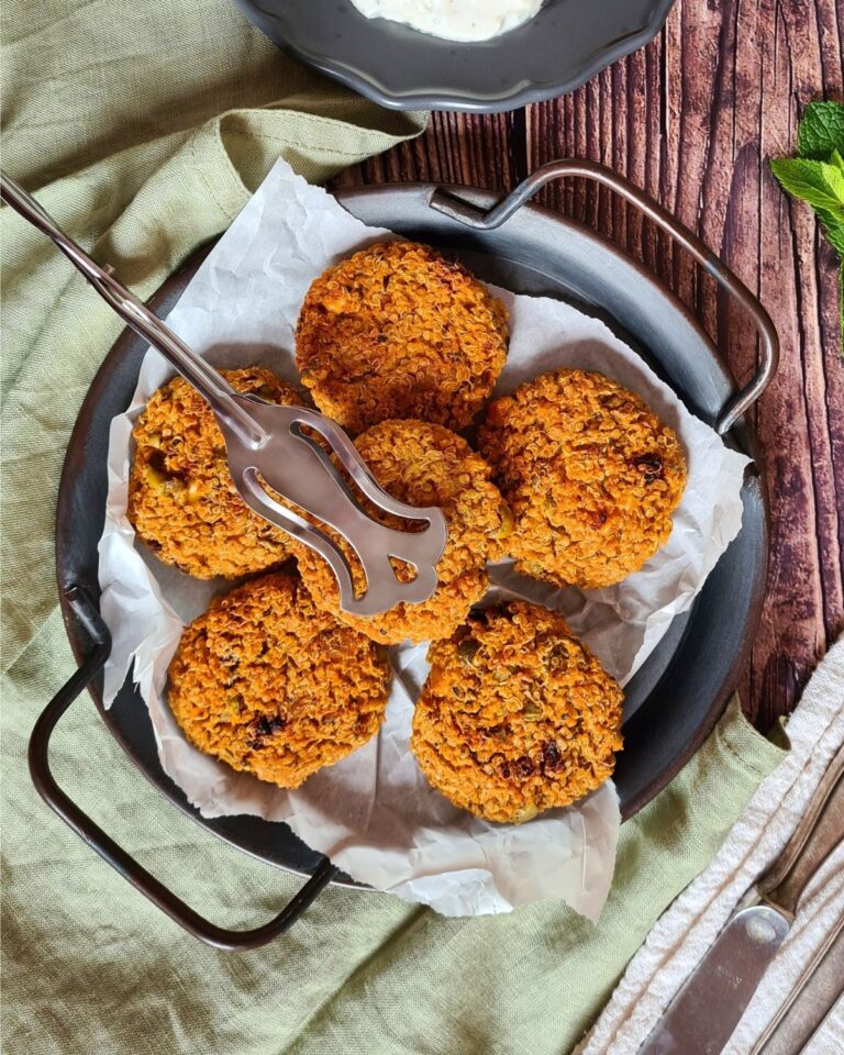 Auf einer runden schwarzen Servierplatte liegen sechs goldbraune Quinoa-Nuggets auf zerknülltem Pergamentpapier. Auf den Bratlingen liegt ein geschlitzter Metallspatel. Die Platte steht auf einem rustikalen Holztisch, unter dem teilweise eine grüne Tischdecke zu sehen ist.