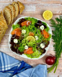 Ein frischer Räucherlachssalat in einer quadratischen weißen Schüssel mit Räucherlachs, Salat, Mozzarellakugeln, roten Zwiebelringen und Zitronenscheiben steht auf einem Holztisch. Um die Schüssel herum liegen Scheiben Mehrkornbrot, eine halbierte Zitrone, frischer Dill, eine rote Zwiebel und eine blau karierte Stoffserviette.