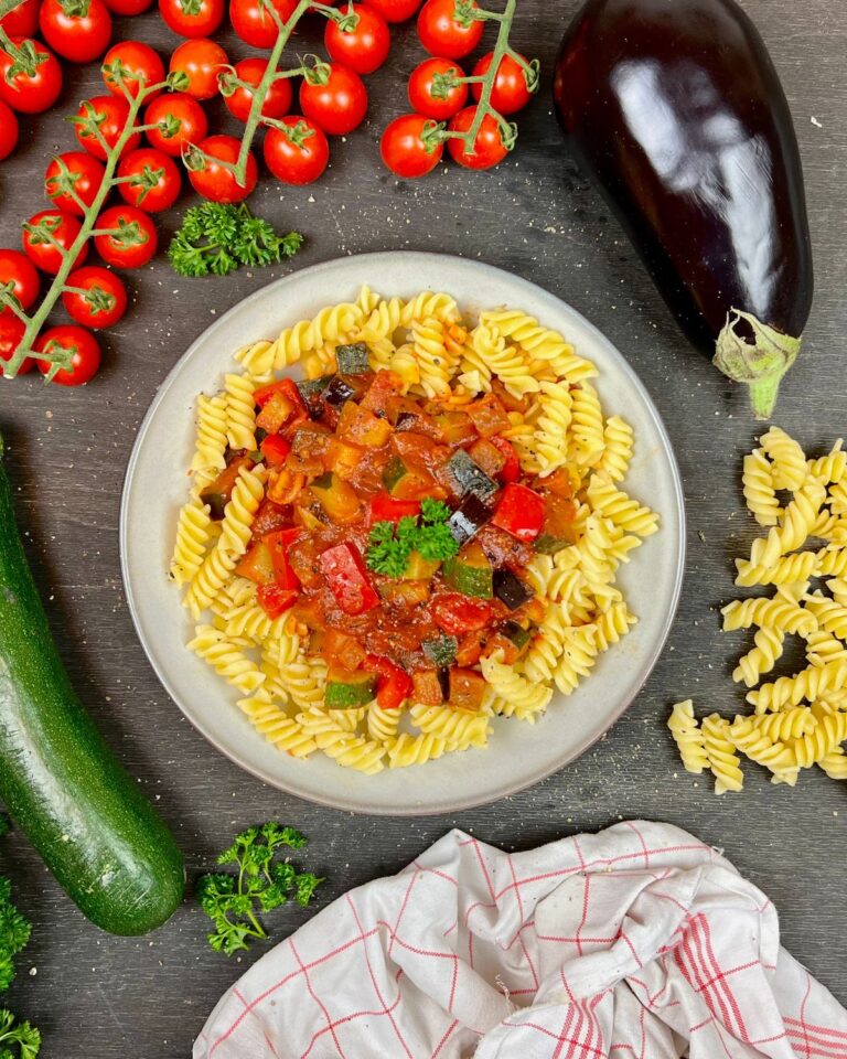 Ein Teller mit Ratatouille-Pasta, garniert mit einer bunten Gemüsesauce, umgeben von frischen Zutaten wie Cherrytomaten, einer Aubergine, einer Zucchini und Petersilie. Außerdem ist ein weiß-rot kariertes Tuch zu sehen. Das Ganze steht auf einer dunklen Oberfläche.