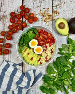 Eine wei&szlig;e Sch&uuml;ssel mit einer Schnellen Couscous-Bowl mit Avocadoscheiben, halbierten gekochten Eiern, Cherrytomaten und Spinat steht auf einer rustikalen Holzoberfl&auml;che.
