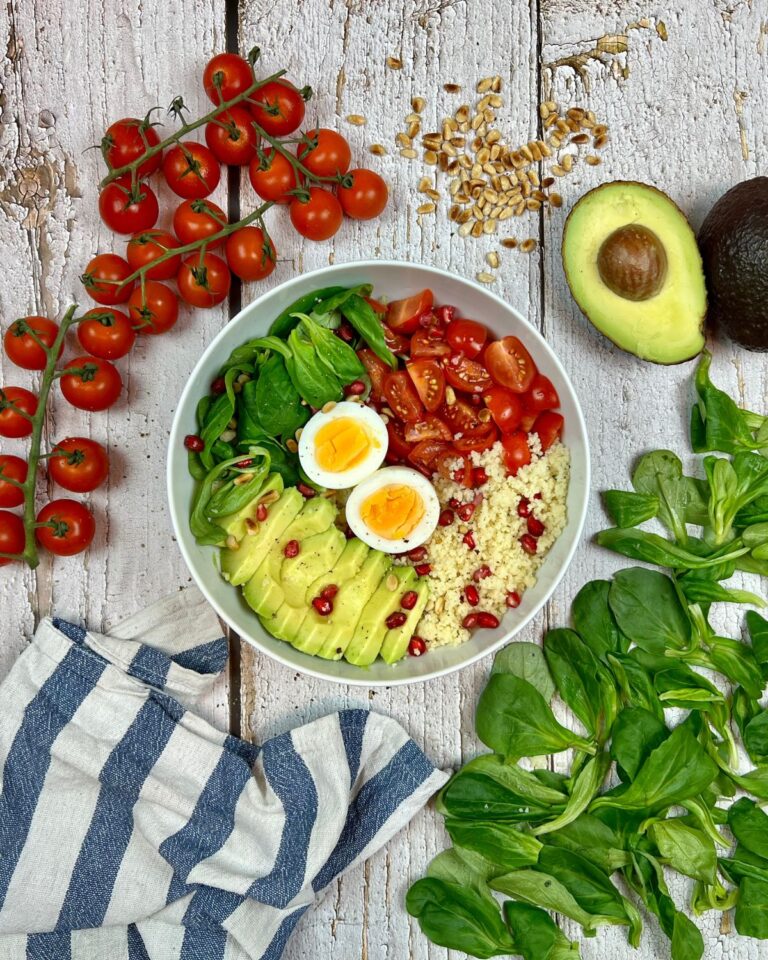 Eine weiße Schüssel mit einer Schnellen Couscous-Bowl mit Avocadoscheiben, halbierten gekochten Eiern, Cherrytomaten und Spinat steht auf einer rustikalen Holzoberfläche.