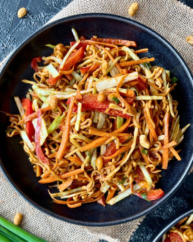 Ein schneller asiatischer Nudelsalat mit geschnittenen Karotten, roten Paprika, Kohl und Erdnüssen. Dieser schnelle asiatische Nudelsalat ist mit einer Erdnuss-Soße beträufelt und ruht auf einer rustikalen Serviette vor einem dunklen Hintergrund.