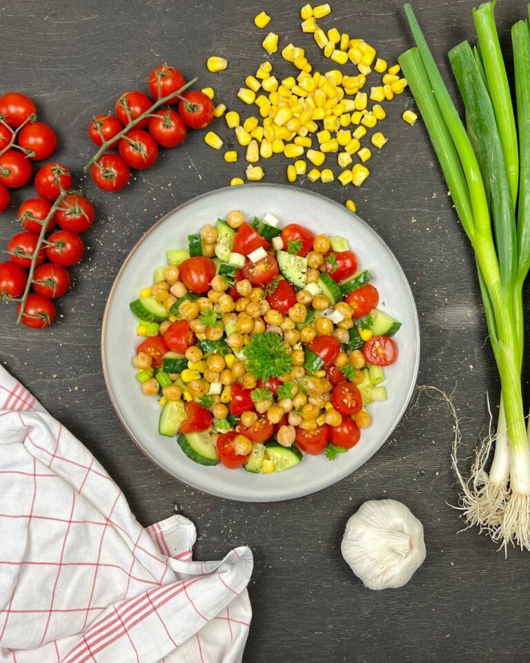 Ein farbenfroher schneller Kichererbsensalat wird auf einem runden weißen Teller serviert und besteht aus Kichererbsen, Gurkenwürfeln, Cherrytomaten und Maiskörnern, garniert mit Petersilie. Um den Teller herum befinden sich frische Zutaten, darunter Cherrytomaten, Frühlingszwiebeln, Maisstücke, Knoblauch und eine karierte Serviette.