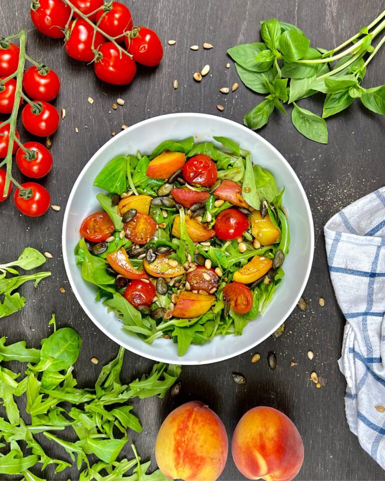 Ein frischer Pfirsich-Salat mit Cherrytomaten und Rucola wird in einer weißen Schüssel serviert, mit saftigen Pfirsichen und knackigen Kürbiskernen.