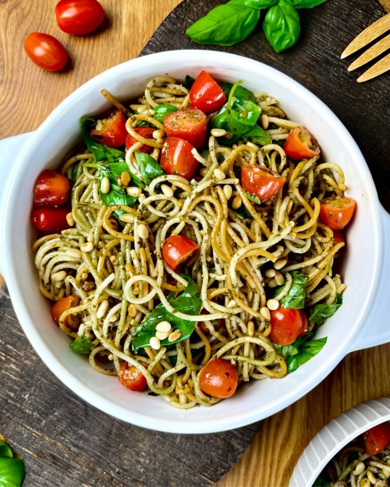 Eine Schüssel Spaghetti-Salat mit Pesto, Cherrytomaten, frischen Basilikumblättern und Pinienkernen steht auf einer Holzoberfläche. Garniert mit zusätzlichem Basilikum und ganzen Tomaten in der Nähe.