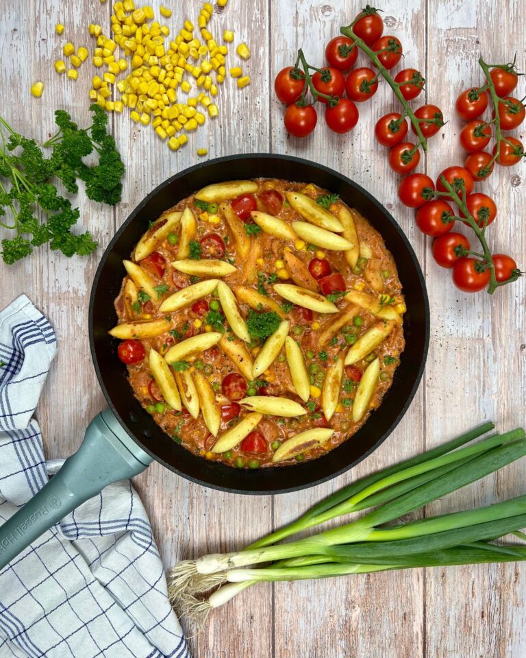 Eine Pfanne Penne-Nudeln vermischt sich mit Kirschtomaten, Mais und Erbsen in einer würzigen Sauce, garniert mit Kräutern. Daneben liegen frische Kirschtomaten am Strauch, Petersilie, Mais, Frühlingszwiebeln und ein kariertes Tuch auf einer Holzoberfläche – eine reizende Anspielung auf den Charme der Schupfnudel-Gemüsepfanne.