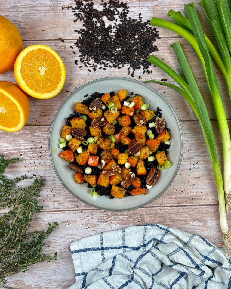 Ein Teller mit schwarzem Reis, Kürbis auf Frühlingszwiebeln steht auf einer Holzoberfläche. Um den Teller herum liegen frische Orangenhälften, wilde Reiskörner, Frühlingszwiebeln, Thymianzweige und ein gestreiftes Küchentuch.