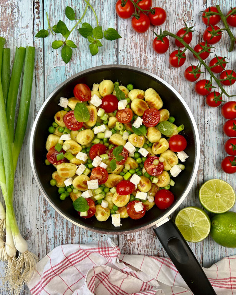 Eine schwarze Pfanne auf rustikaler Holzoberfläche enthält eine sommerliche Gnocchi-Pfanne mit Cherrytomaten, Erbsen und Fetakäsewürfeln, garniert mit frischen Basilikumblättern. Um die Pfanne herum liegen frische Frühlingszwiebeln, Kirschtomaten am Strauch, Limettenhälften und ein rot-weißes Tuch.