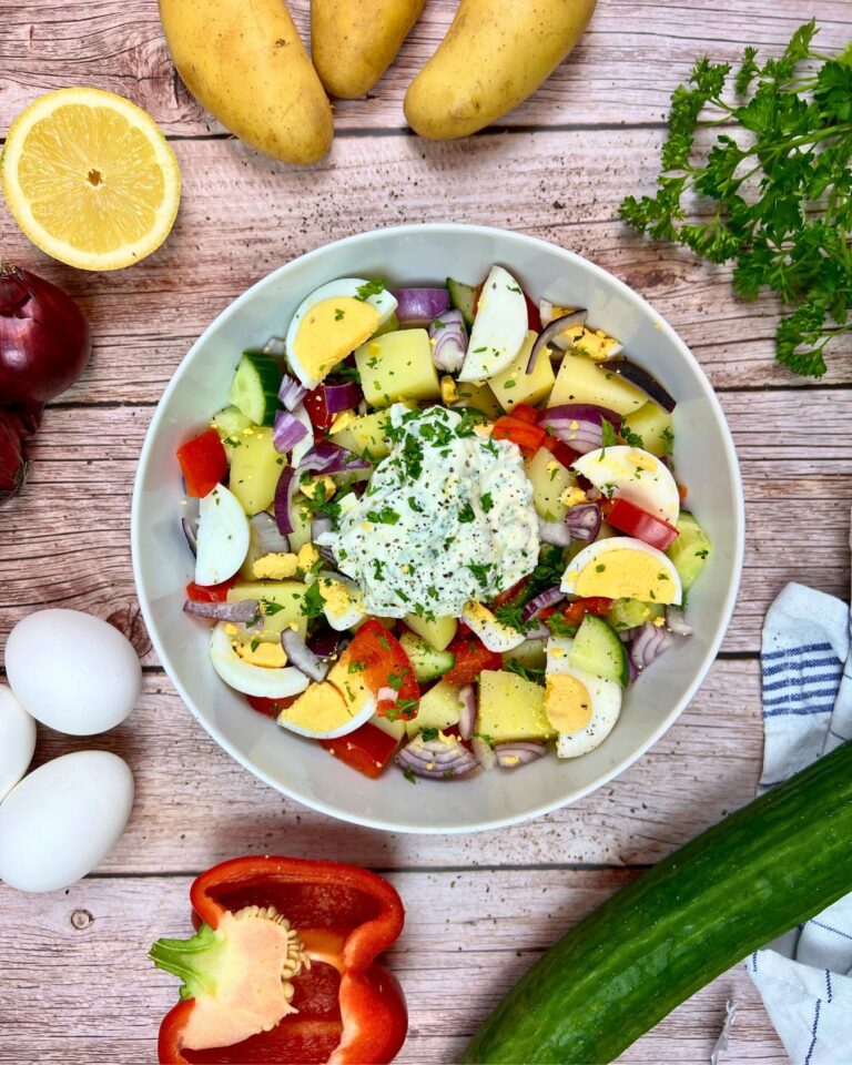 Auf einem Holztisch steht eine Schüssel sommerlicher Kartoffelsalat mit Kartoffeln, hartgekochten Eiern, roten Zwiebeln, Gurken und Paprika, getoppt mit ein wenig Frischkäse.