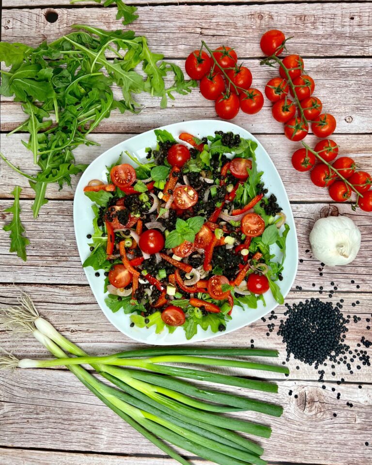 Eine Schüssel sommerlicher Linsensalat mit Cherrytomaten, schwarzen Beluga-Linsen und Blattgemüse steht auf einer Holzfläche.