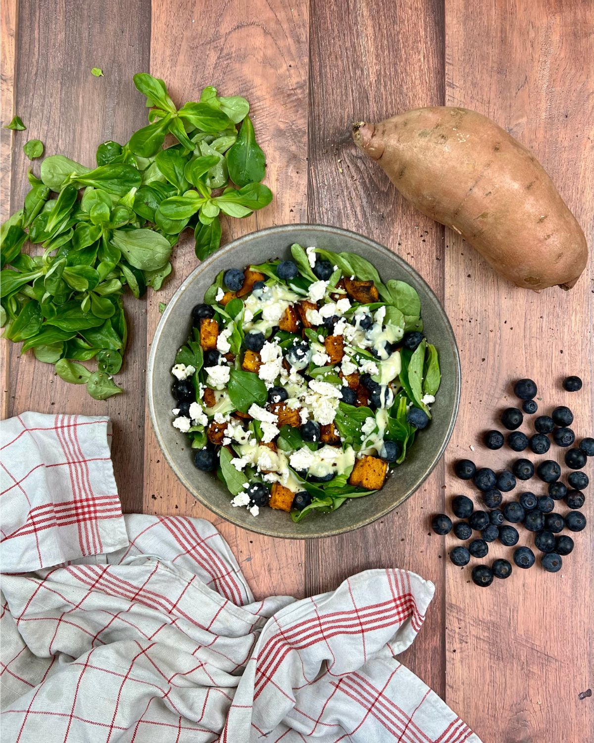 Eine Salatschüssel auf einer Holzoberfläche, mit Feldsalat, gerösteten Süßkartoffelstücken, Blaubeeren, Fetakäse und einem cremigen Dressing. Um die Schüssel herum befinden sich eine Süßkartoffel, weitere Blaubeeren und ein kariertes Tuch.