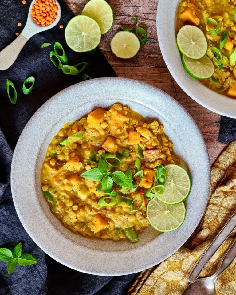 Eine herzhafte Schüssel Süßkartoffel-Linsen-Curry, garniert mit frischen Basilikumblättern und Limettenscheiben. Ein Holztisch mit einer weiteren Schüssel Eintopf, einem Löffel, einer gelben Stoffserviette, geschnittenen Frühlingszwiebeln und einem Löffel roten Linsen im Hintergrund.