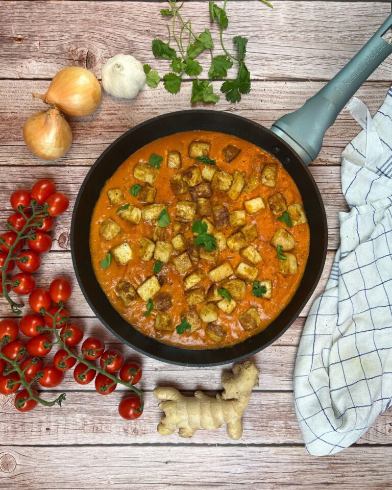 Eine Pfanne mit cremigem Tofu Tikka Masala, belegt mit gerösteten Tofustücken und garniert mit frischem Koriander. Um die Pfanne herum liegen Zwiebeln, Knoblauch, Kirschtomaten, Ingwer und ein kariertes Tuch auf einer Holzoberfläche.