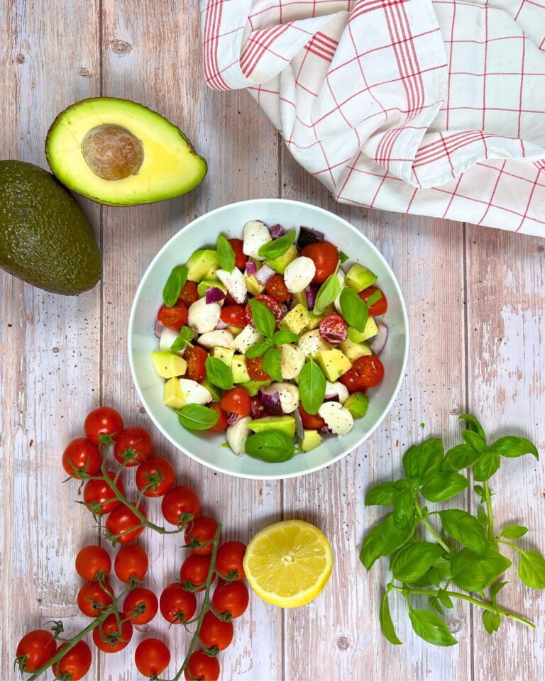 Eine Schüssel Tomaten-Avocado-Salat mit Mozzarellakugeln, Avocado, Cherrytomaten und Basilikum steht auf einem Holztisch. Um ihn herum sind ganze Avocados, eine Reihe Cherrytomaten, eine halbe Zitrone, Basilikumblätter und ein Tuch für zusätzliches Ambiente.