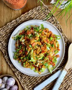 Ein Teller mit veganem Linsensalat aus roten Linsen steht auf einer geflochtenen Matte. Dieser farbenfrohe vegane Linsensalat enthält gehacktes Grün, rote Cherrytomaten und anderes kleines Gemüse. Um den Teller herum stehen ein Glas mit roten Linsen, Knoblauchknollen, ein weißer Blumentopf und Holzutensilien.